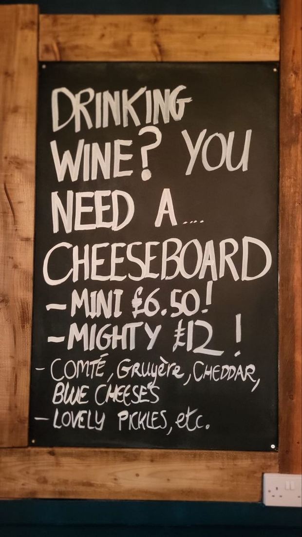 The pub's chalkboard: Drinking wine? You need a… cheeseboard. Mini £6.50! Mighty £12! Comté, Gruyère, Cheddar, blue cheeses. Lovely pickles, etc.