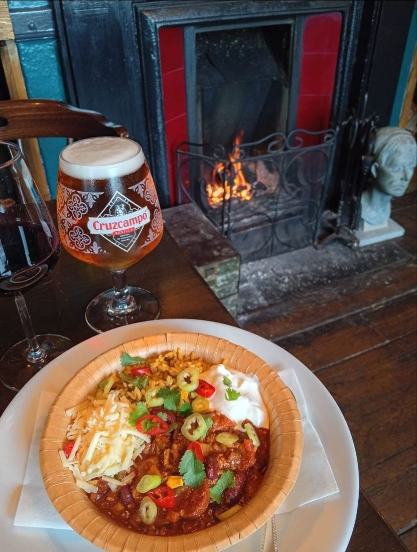 A bowl of home cooking with a beer and a glass of red wine in front of the lit fire.