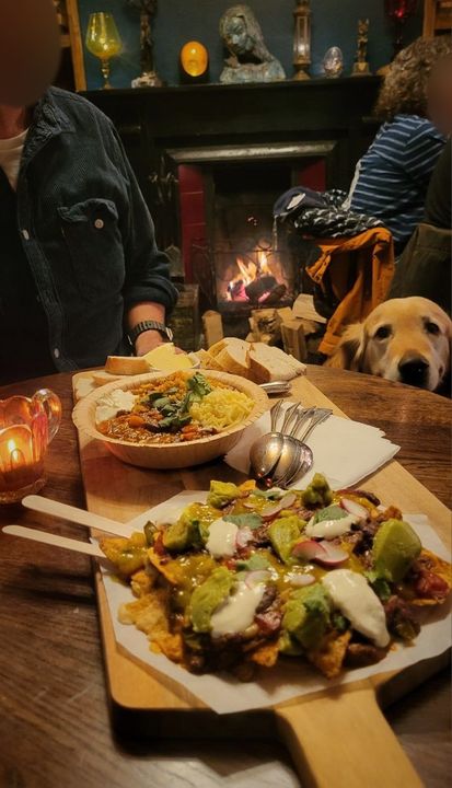 Nachos and a bowl of chilli with rice on a long wooden board by the lit fire, with bread and butter alongside. A golden retriever peers over the edge of the table at the food.