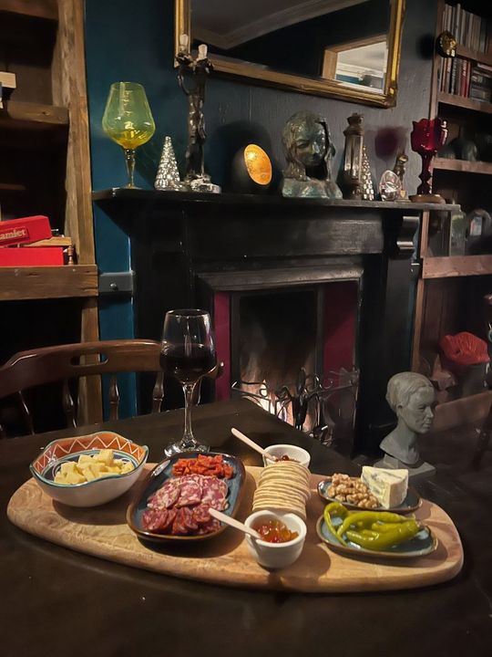 A sharing board by the lit fire with cheese, cured meats, crackers, walnuts, blue cheese, pickled green chillies and chutney, beside a glass of red wine. Busts and curiosities line the mantel under a gilt mirror.