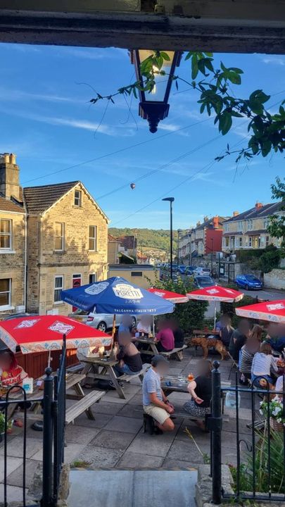 The view from the doorway under a lantern on a summer evening. People sit at picnic tables under red and blue parasols with a dog among them, and the Bath stone houses and green hills beyond glow in the low sun. A hot-air balloon floats in the blue sky.