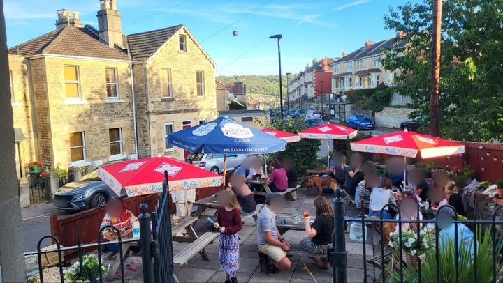 The beer garden full of people of all ages on a sunny evening, at picnic tables under red and blue parasols, with a dog among them. Bath stone houses and green hills stand behind, and a hot-air balloon floats in the blue sky.