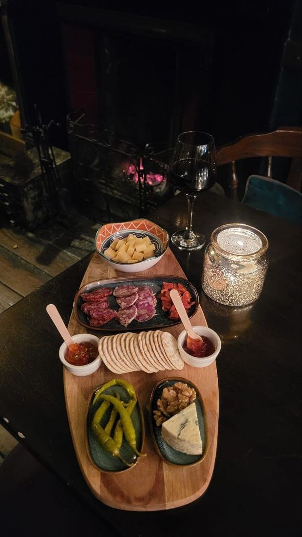 A long wooden sharing board with a glass of red wine and a candle in front of the glowing fire.