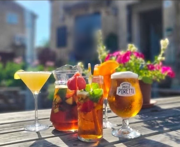Drinks on a wooden table in the sun with pink flowers behind. There&rsquo;s a cocktail with lime, a jug and a tall glass full of fruit, an orange spritz and a beer.